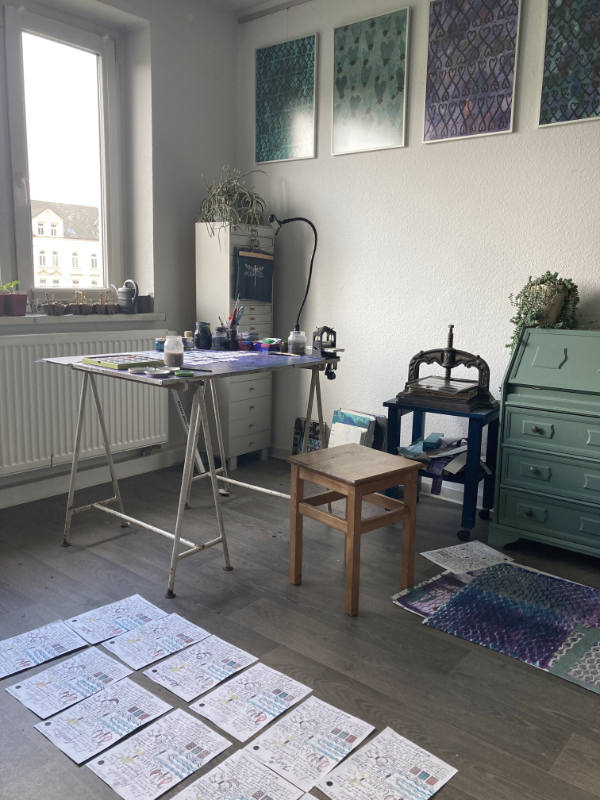 Blick in das Tatelier mit Lila Tisch, MiniGalerie oben an der Wand und Buchpresse im Hintergrund. Auf dem Fußboden trocknen kolorierte Bildbriefe